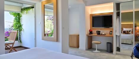 A modern hotel room with a large bed, a TV, a desk with a chair, and a sliding door leading to a balcony with plants.
