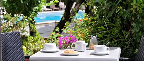 A table set for two with white cups, a plate of cookies, and a pitcher, surrounded by lush greenery and a pool in the background.