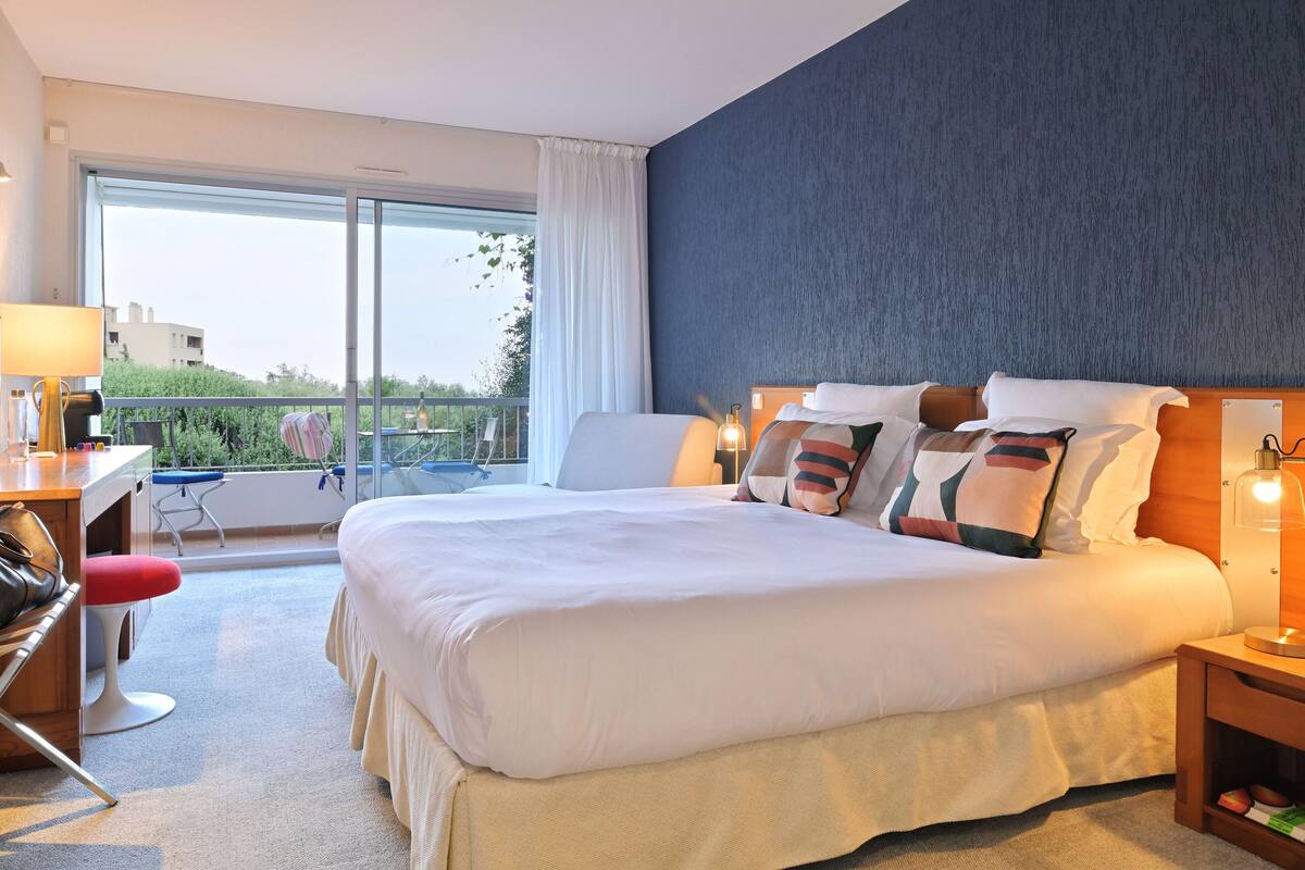 Habitación de hotel con una cama grande, un escritorio con silla, una mesita y una mesita de noche con lámpara.