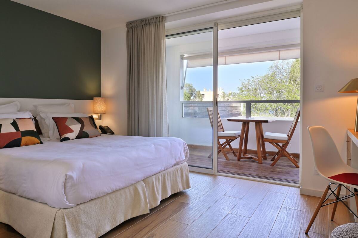 Habitación de hotel con una cama grande, un balcón con mesa y sillas, y vistas al exterior.