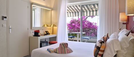 A hotel room with a bed, a desk with a lamp, a coffee maker, a mirror, and a view of purple flowers through the window.