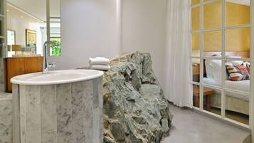 A bathroom with a marble sink, a large rock feature, and a view of a bedroom with a bed and a window.
