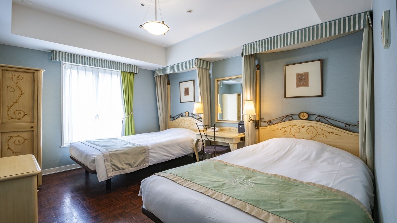 Quarto de hotel com duas camas, uma escrivaninha, uma cadeira, um guarda-roupa e uma janela com cortinas.