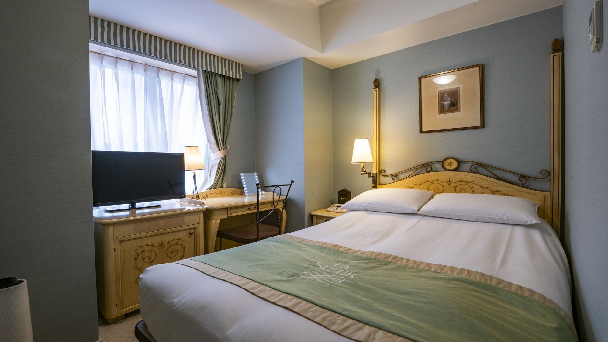 Quarto de hotel com cama, escrivaninha com televisão, cadeira, mesa de cabeceira e luminária.