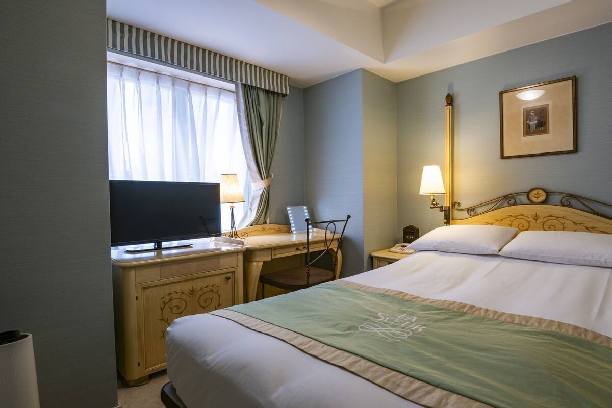 Habitación de hotel con cama, escritorio con televisor, silla, mesita de noche y lámpara.