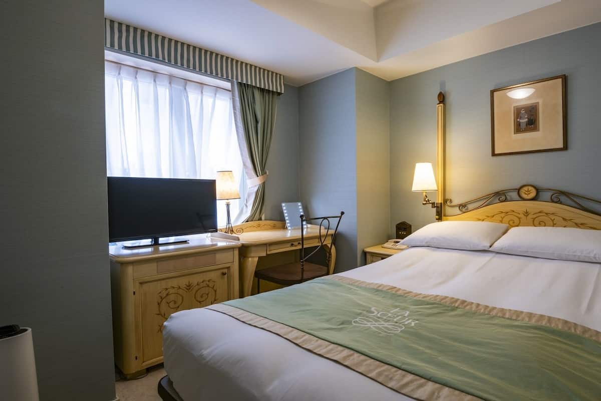 A hotel room with a bed, a desk with a TV, a chair, a bedside table, and a lamp.
