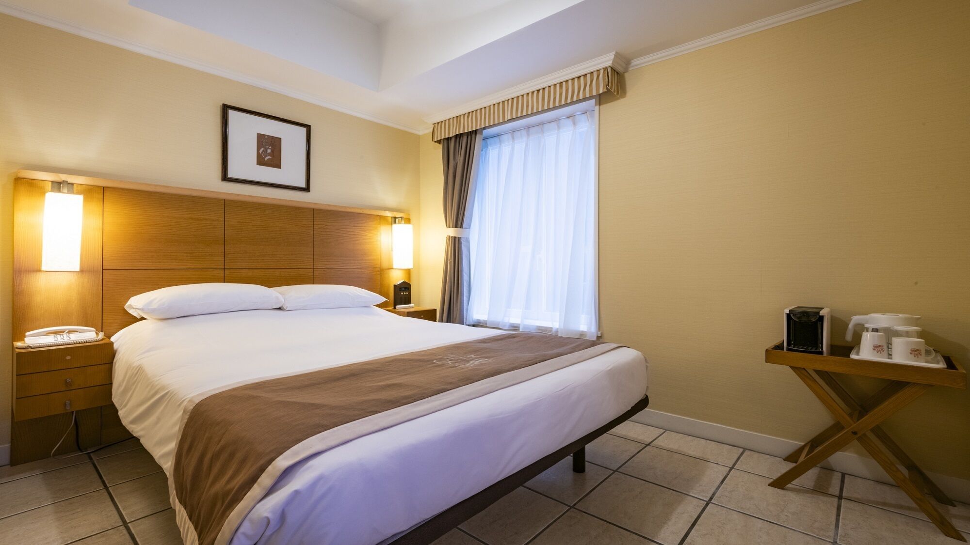 Quarto de hotel com cama, abajures, telefone, cafeteira e um quadro na parede.