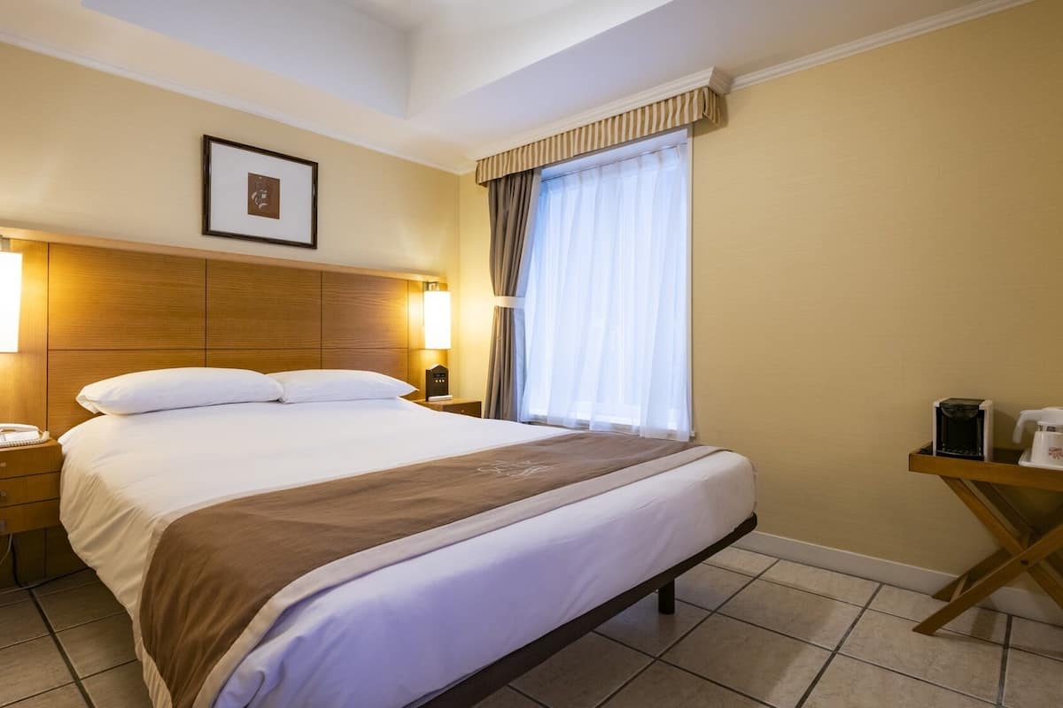 Habitación de hotel con cama, lámparas de noche, teléfono, cafetera y un cuadro en la pared.