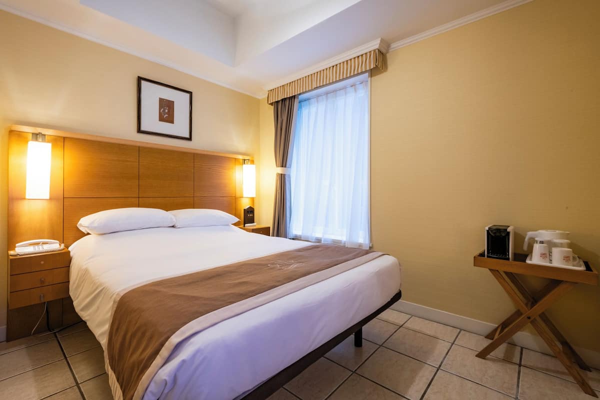Habitación de hotel con una cama grande, lámparas de noche, mesita de noche con un teléfono, una pequeña mesa con cafetera y una ventana con cortinas.