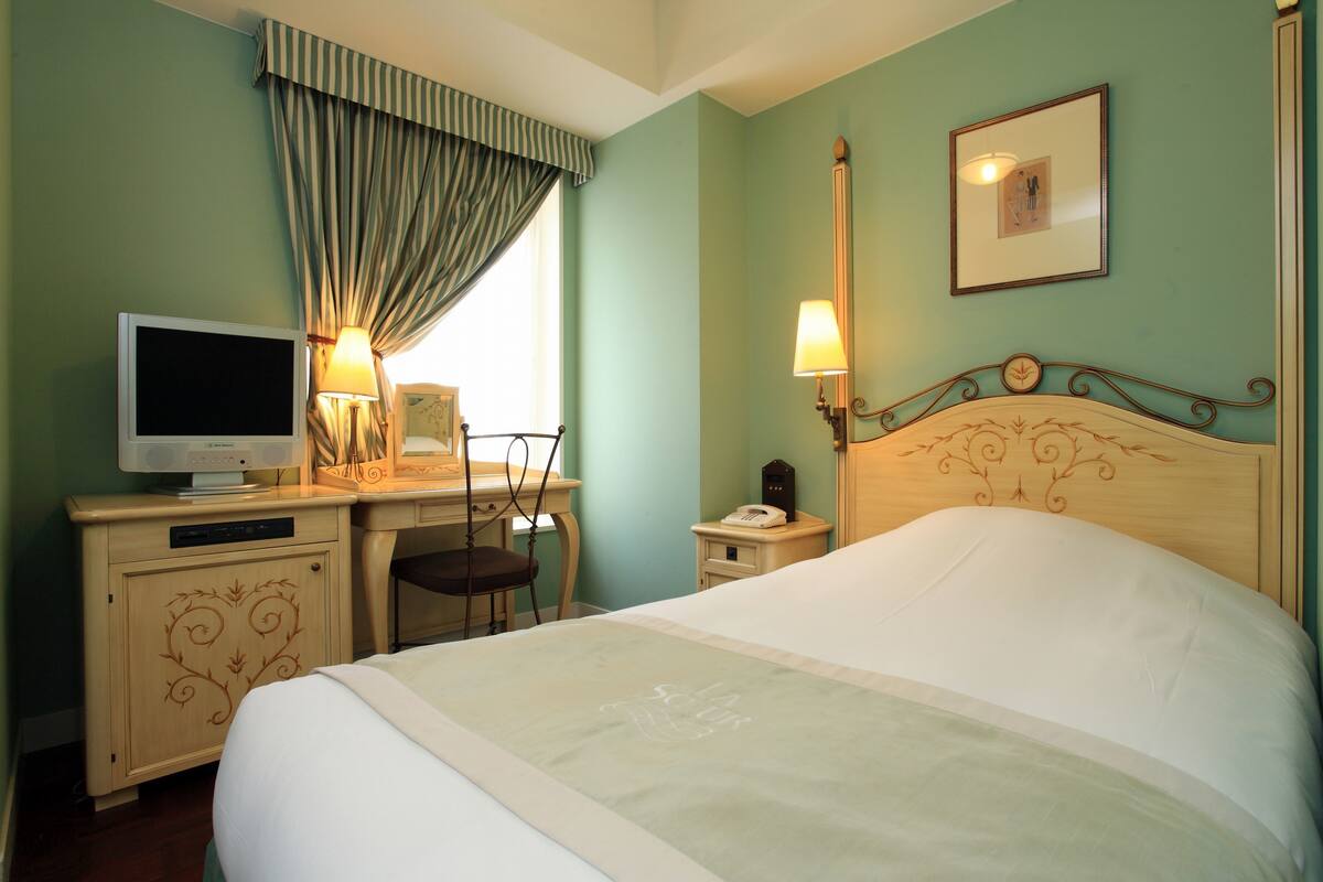 Habitación de hotel con cama, escritorio con computadora, silla, lámpara y mesita de noche con teléfono.
