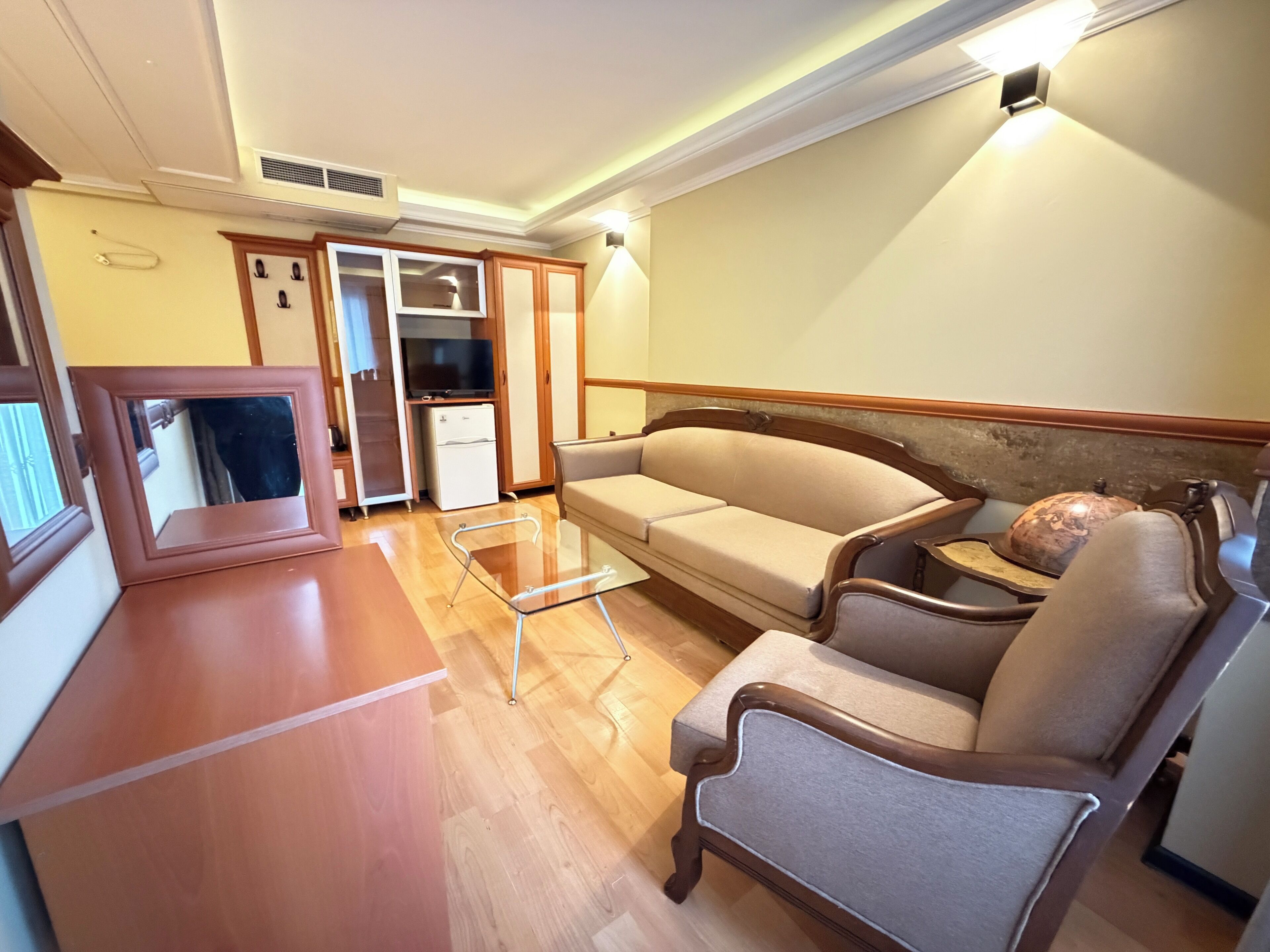 A compact living area with a sofa, armchairs, a glass-top coffee table, a television, and a minibar.