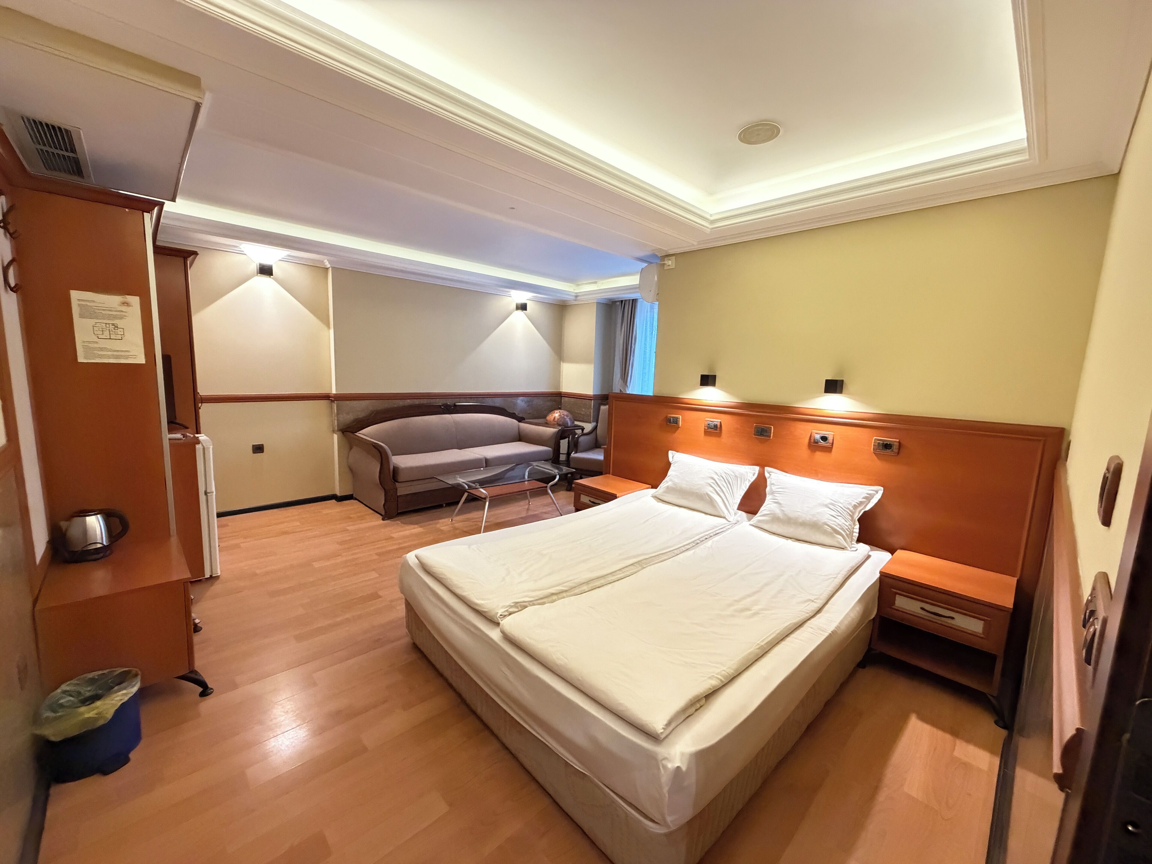 A hotel room with a large bed, a wooden nightstand, a desk with a chair, a sofa, and a wardrobe.