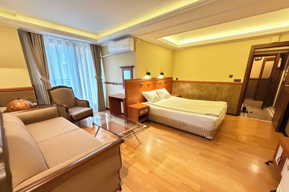 A hotel room with a bed, a sofa, a desk, and an air conditioning unit.