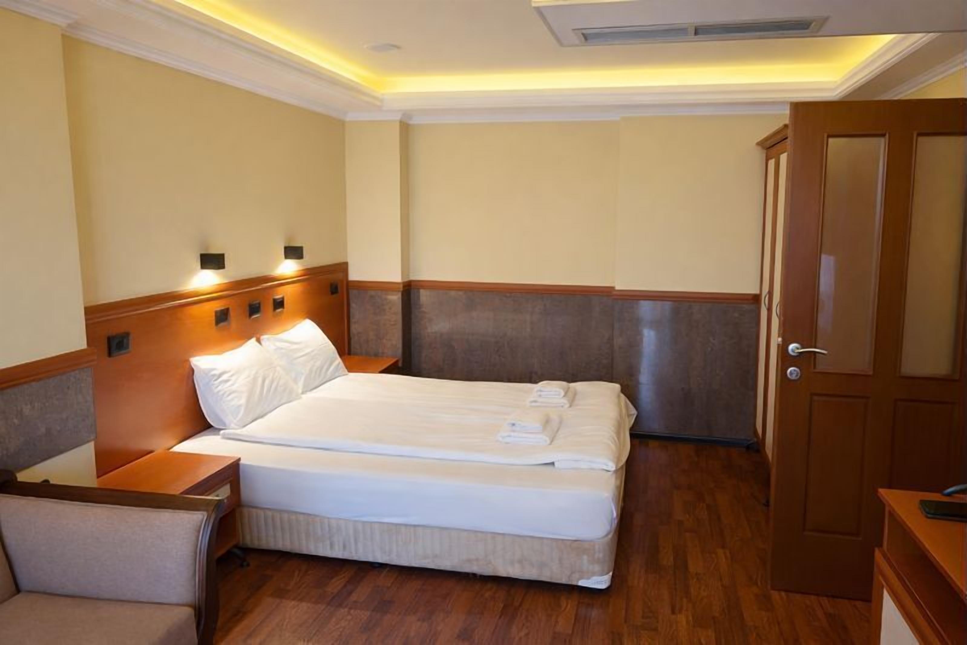 Habitación de hotel con una cama doble, cabecera de madera, mesita de noche, una silla y una puerta.