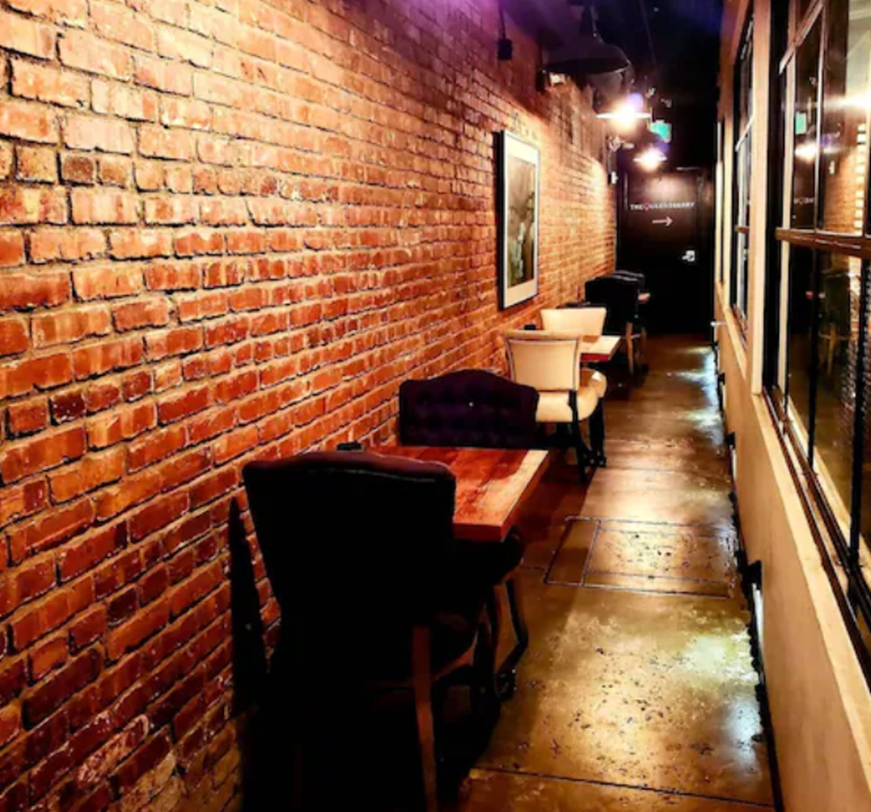 A narrow corridor with brick walls, a row of tables and chairs, and a framed picture on the wall.
