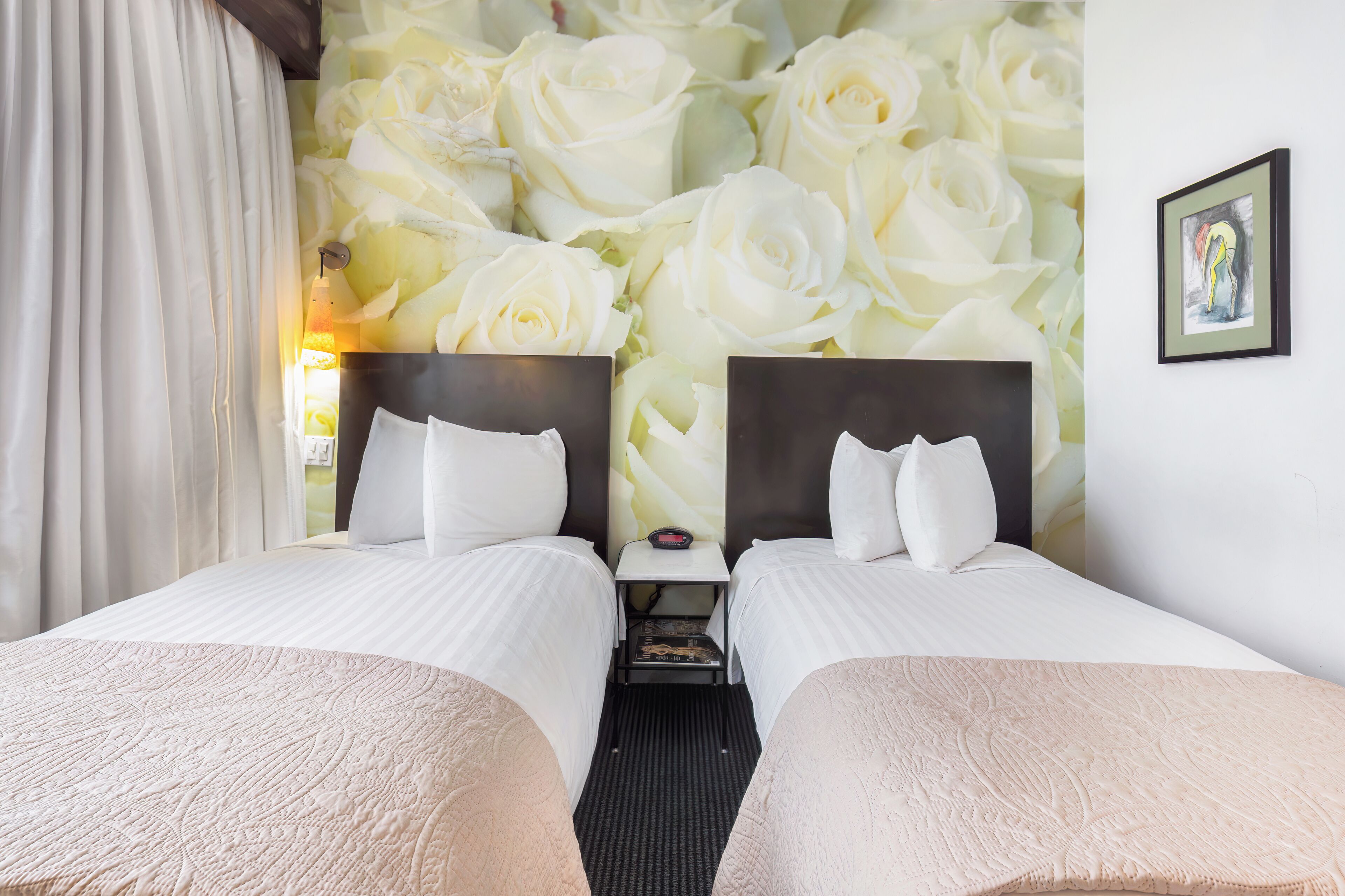 A hotel room with two beds, a nightstand, a wall with a floral wallpaper, a framed picture, and a window with curtains.