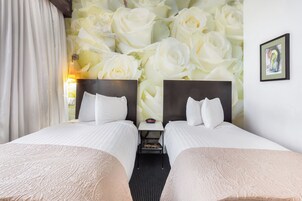 A hotel room with two beds, a nightstand, a wall with a floral wallpaper, a framed picture, and a window with curtains.