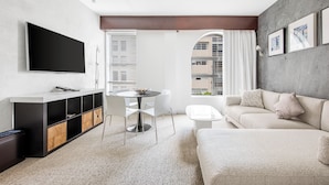 A modern living room with a flat-screen TV, a round dining table, white chairs, a beige sofa, and a built-in entertainment unit. - The O Hotel, Trademark Collection by Wyndham (Los Angeles)