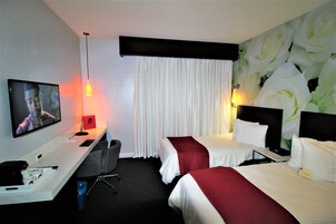 A hotel room with two beds, a desk with a TV, and a large floral wallpaper.