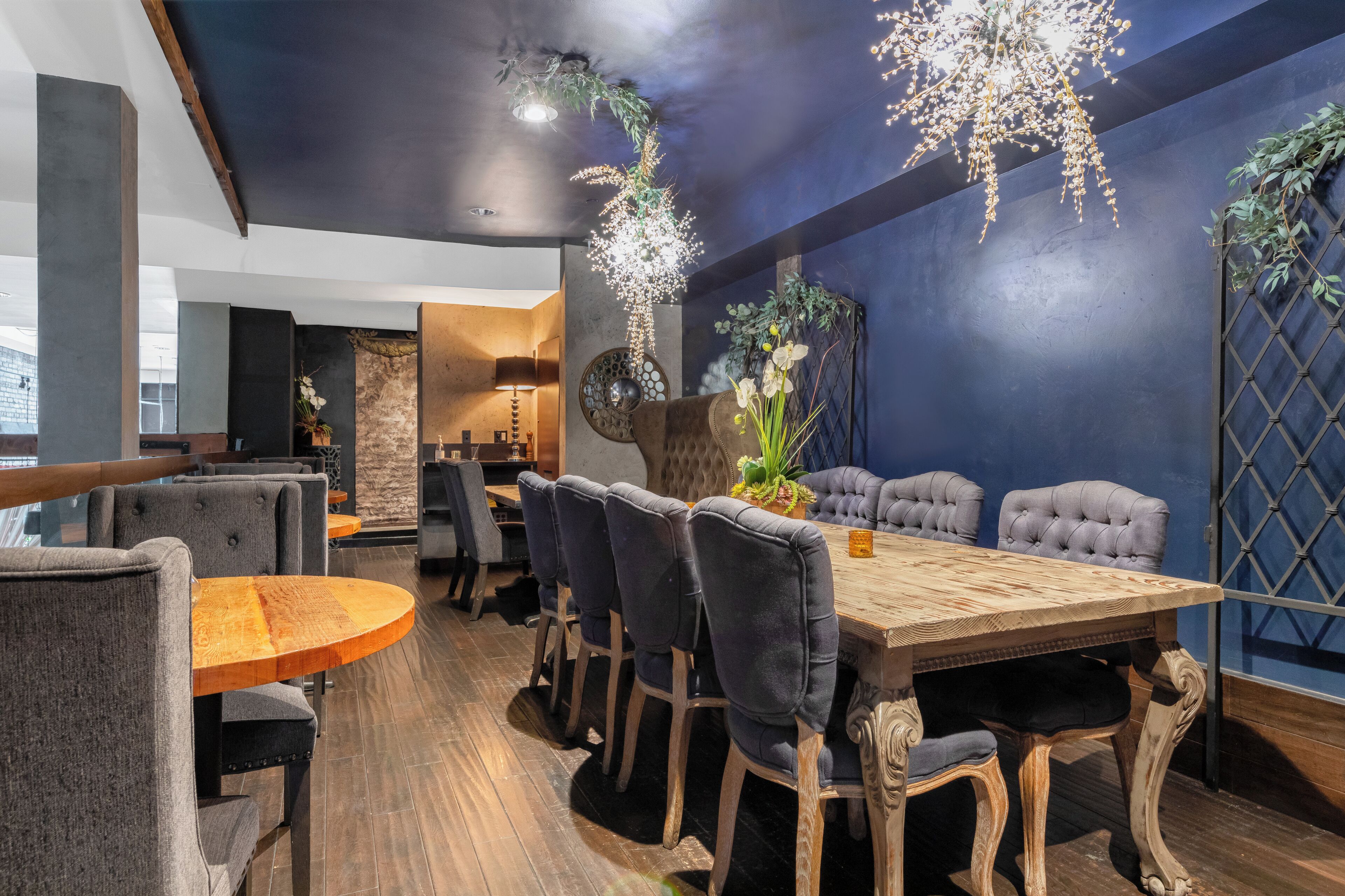 A dining area with a wooden table and upholstered chairs, a chandelier, and decorative plants.