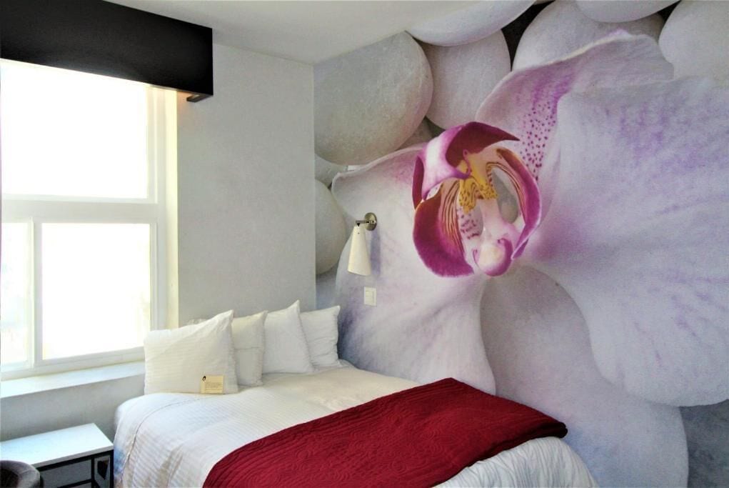 Un dormitorio con un gran mural de orquídeas en la pared, una cama con ropa blanca, un plaid rojo y una lámpara de noche.