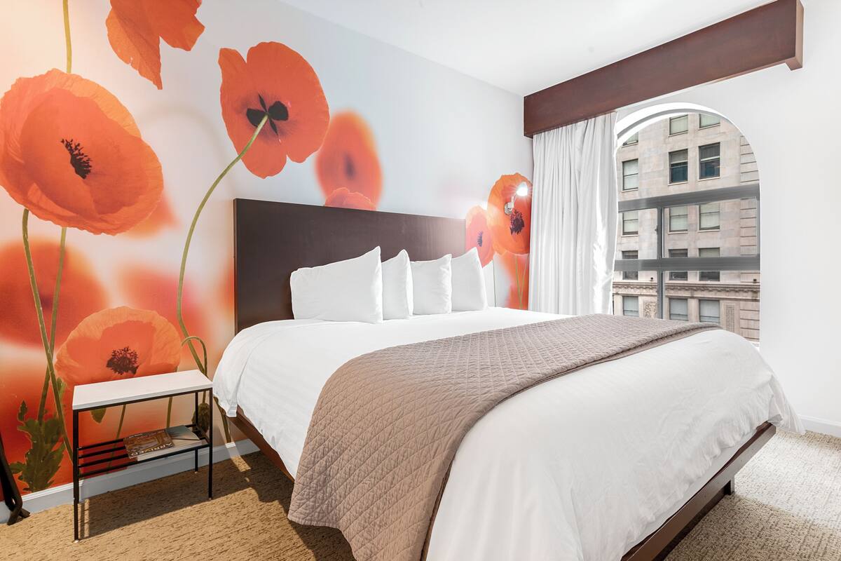 Camera d'albergo con un letto grande, biancheria bianca, un murale con papaveri, una finestra con tende e vista su un edificio.
