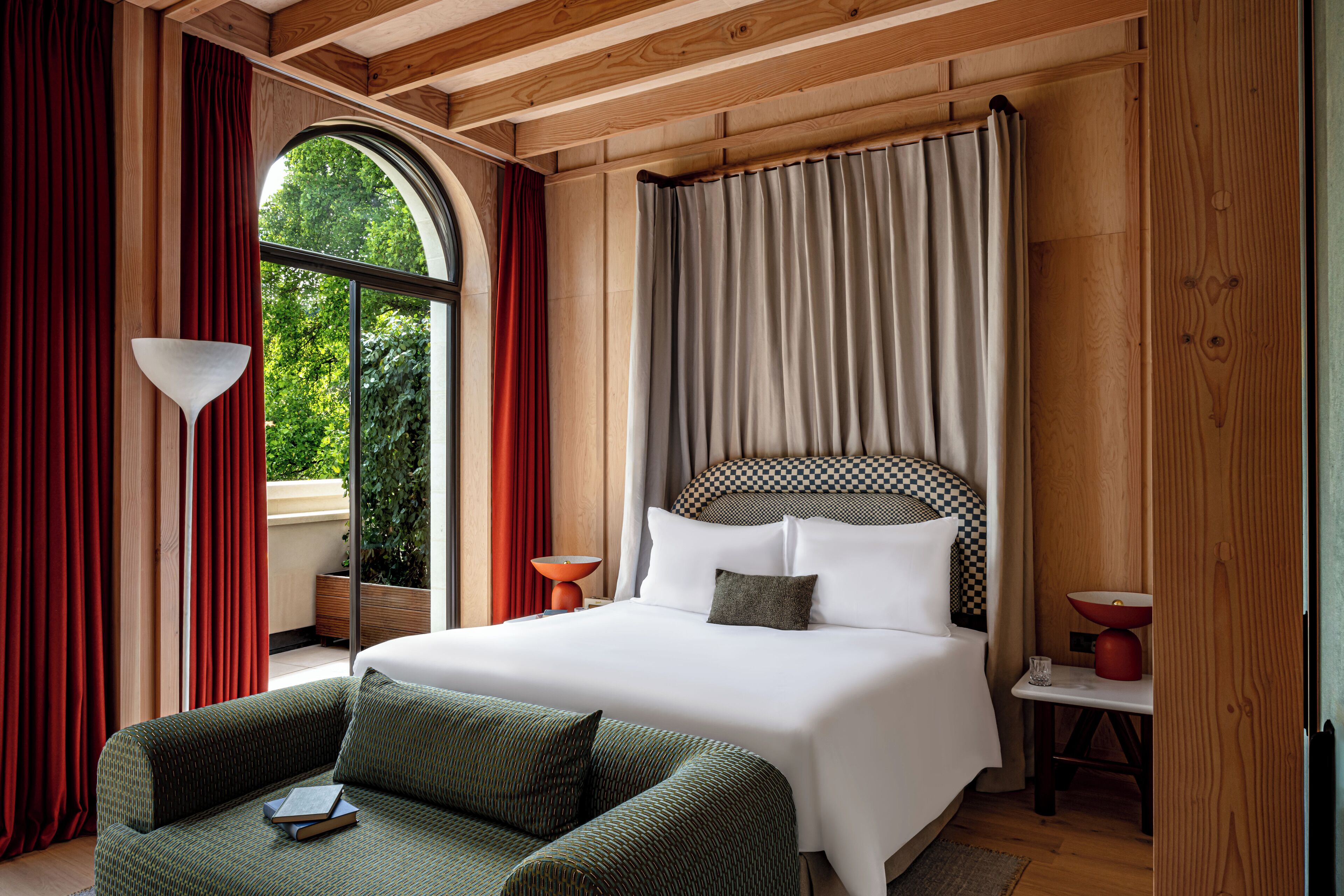 A bedroom with a large bed, a sofa, a window with curtains, and a view of greenery outside.