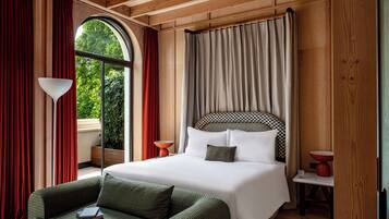 A bedroom with a large bed, a sofa, a window with curtains, and a view of greenery outside.