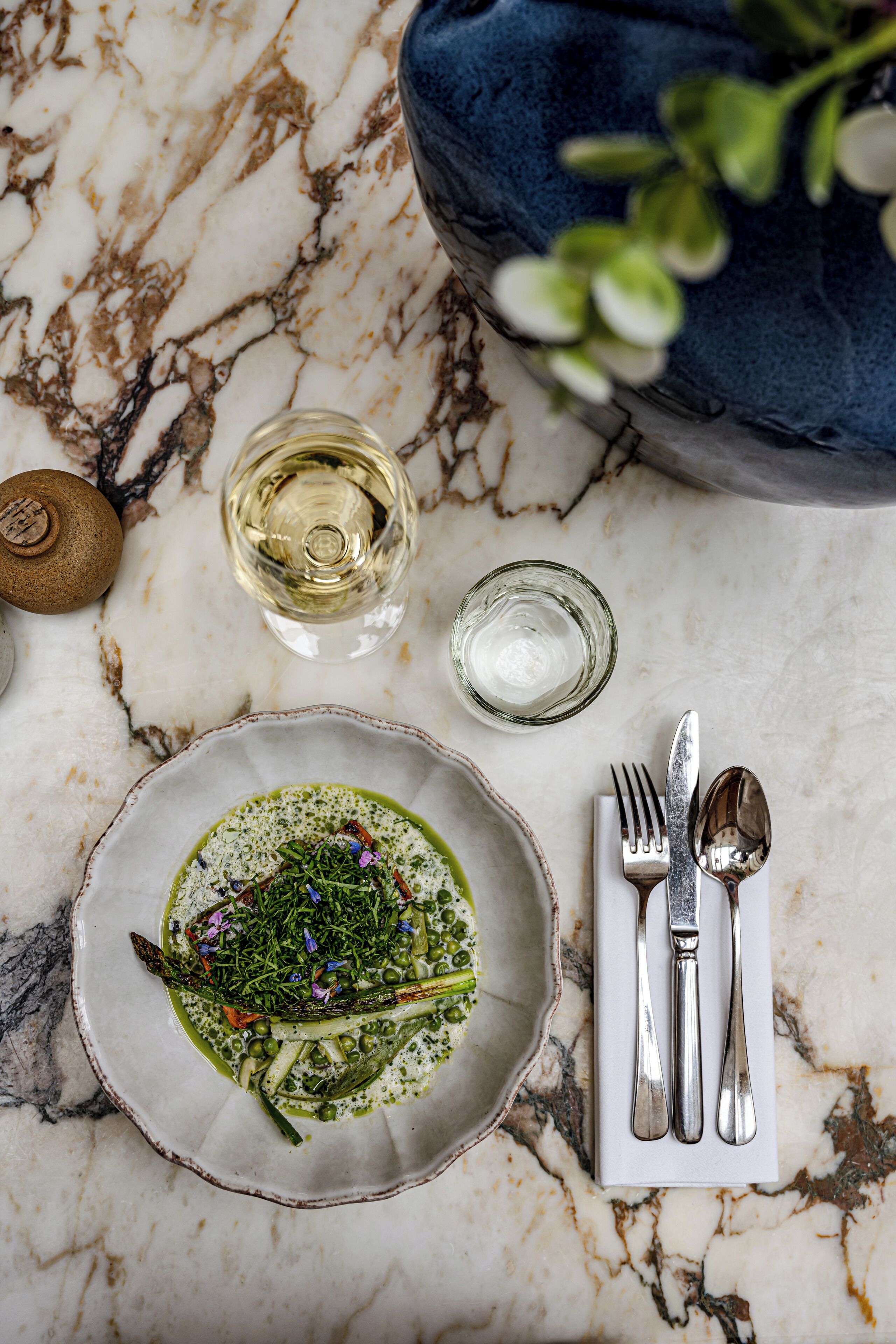 A dining table with a plate of food, a glass of white wine, and cutlery on a marble surface.