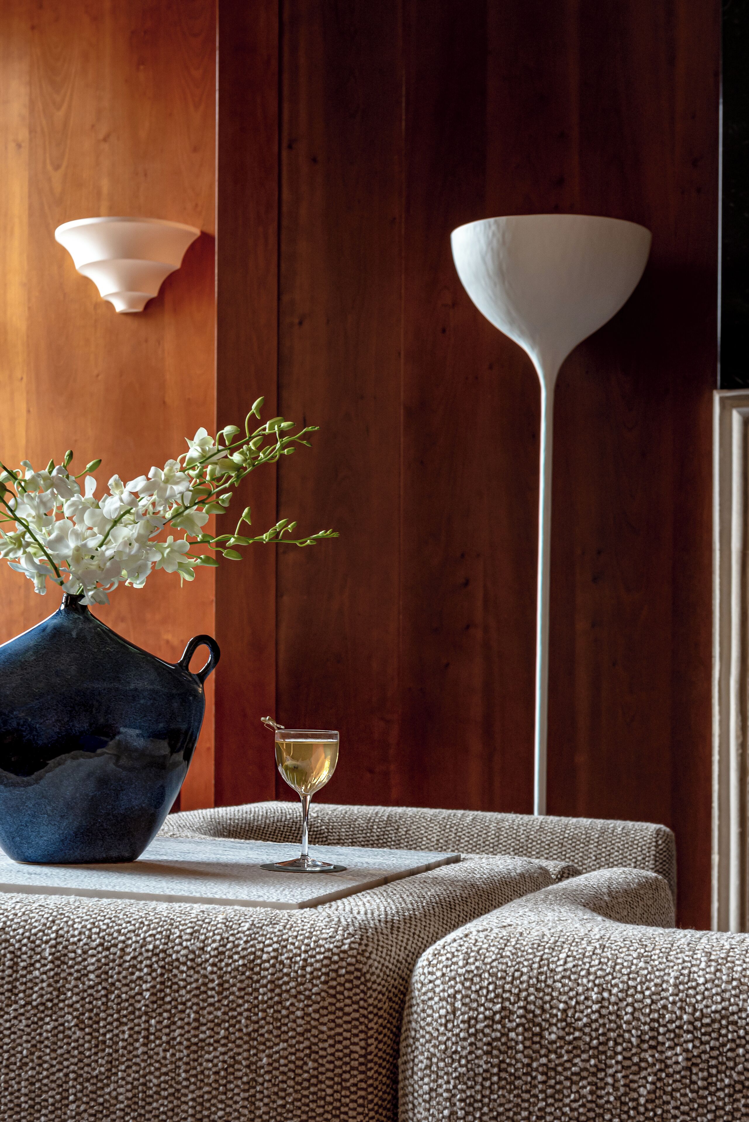 A room with a wooden wall, a blue vase with white flowers, a glass of white wine, and a white floor lamp.