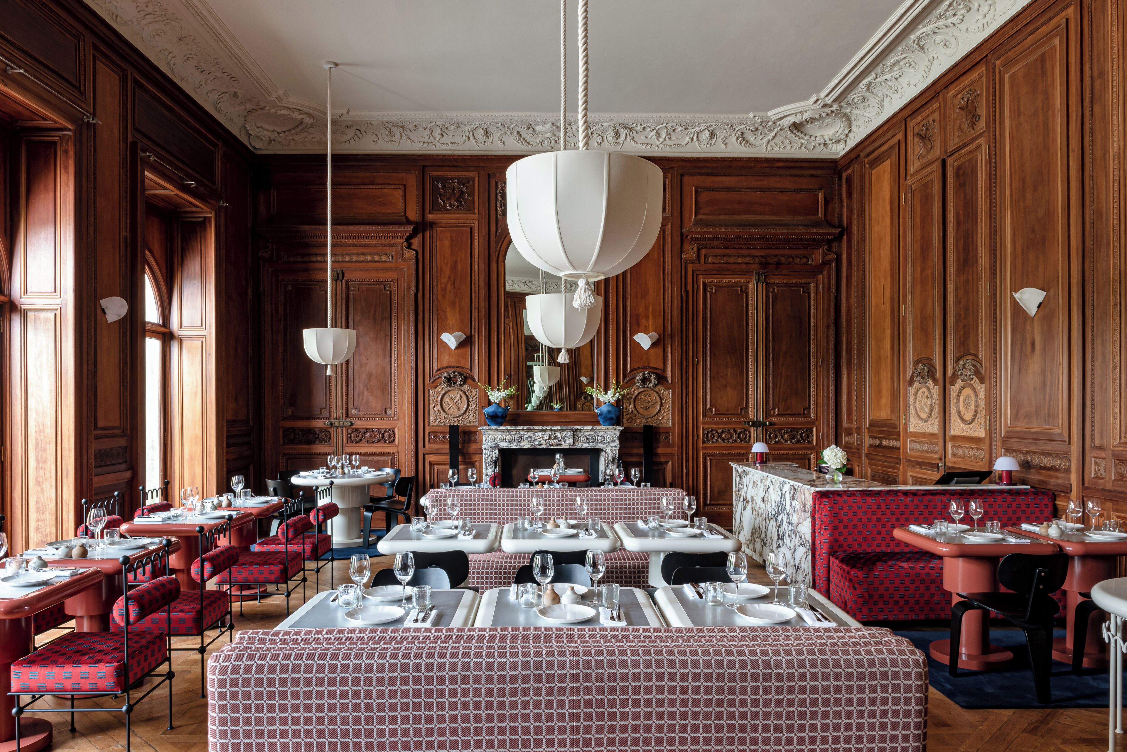 A formal dining room with wooden paneling, marble tables, and red upholstered chairs.