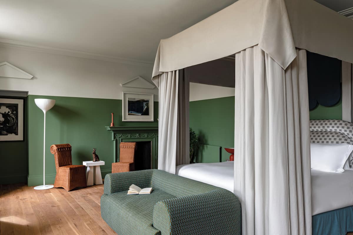 A bedroom with a four-poster bed, a green sofa, a small table, and a fireplace.