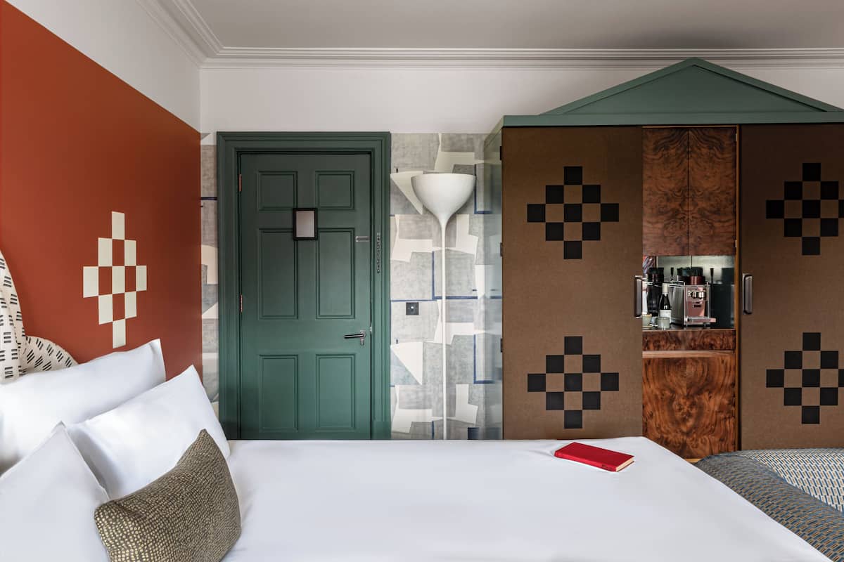 A hotel room with a bed, a green door, a patterned wall, and a wardrobe with a mirror.