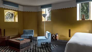 A hotel room with a checkered rug, a bed, a blue patterned chair, a wicker side table, and a floor lamp.