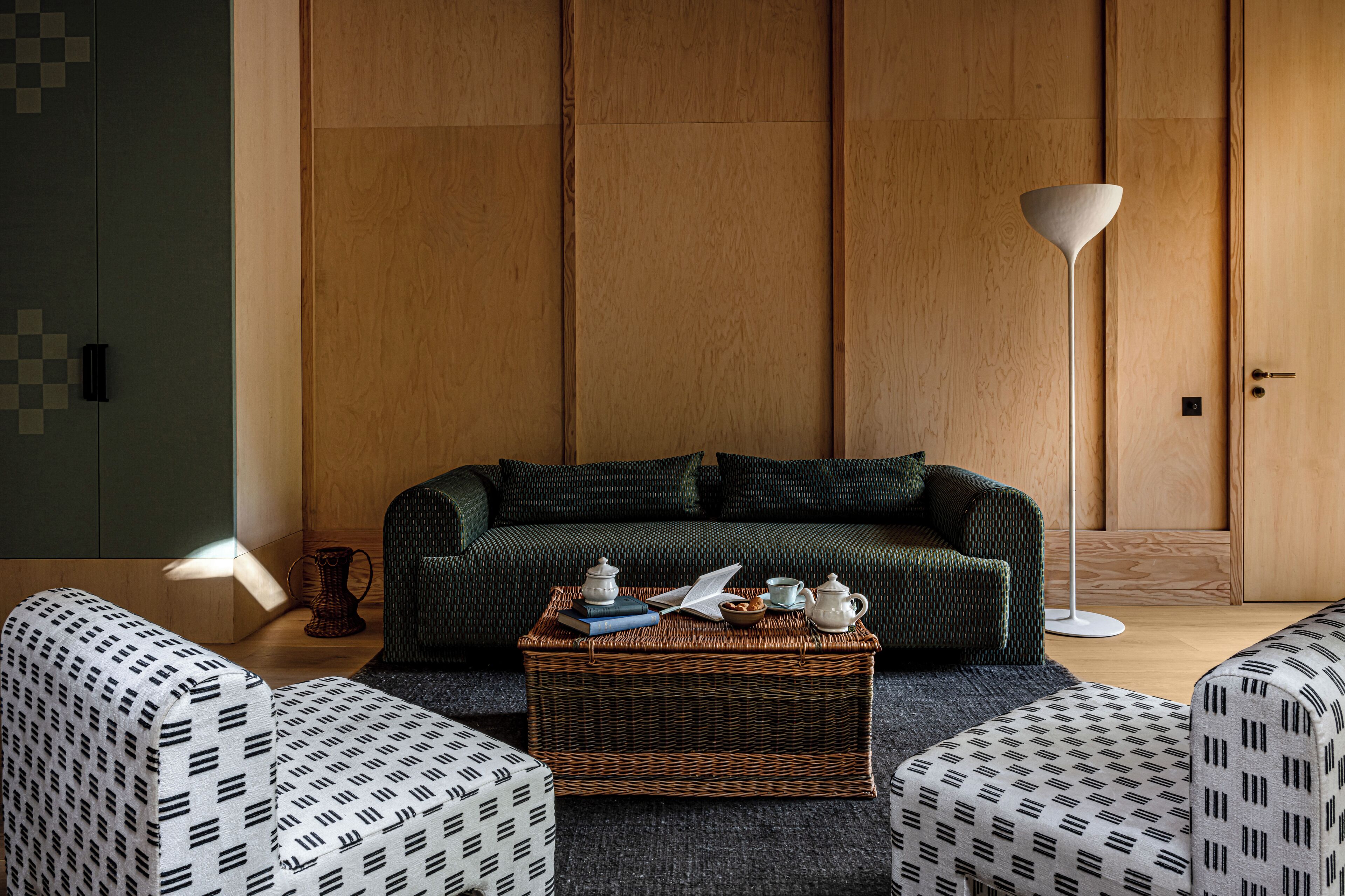 A living room with a sofa, two patterned armchairs, a coffee table with books and teapots, a floor lamp, and wooden panel walls.