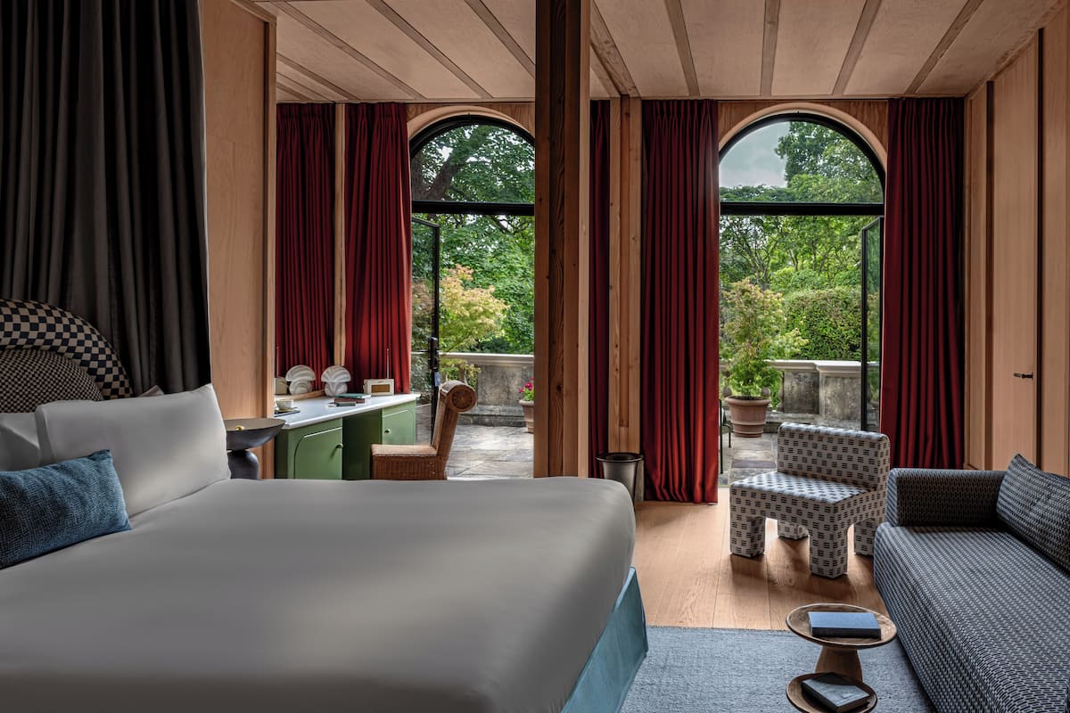 A hotel room with a large bed, a sofa, a desk, and two chairs. There are large windows with curtains and a view of greenery outside.