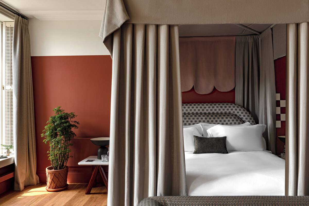 A hotel room with a bed, curtains, a potted plant, a table, and a window with blinds.