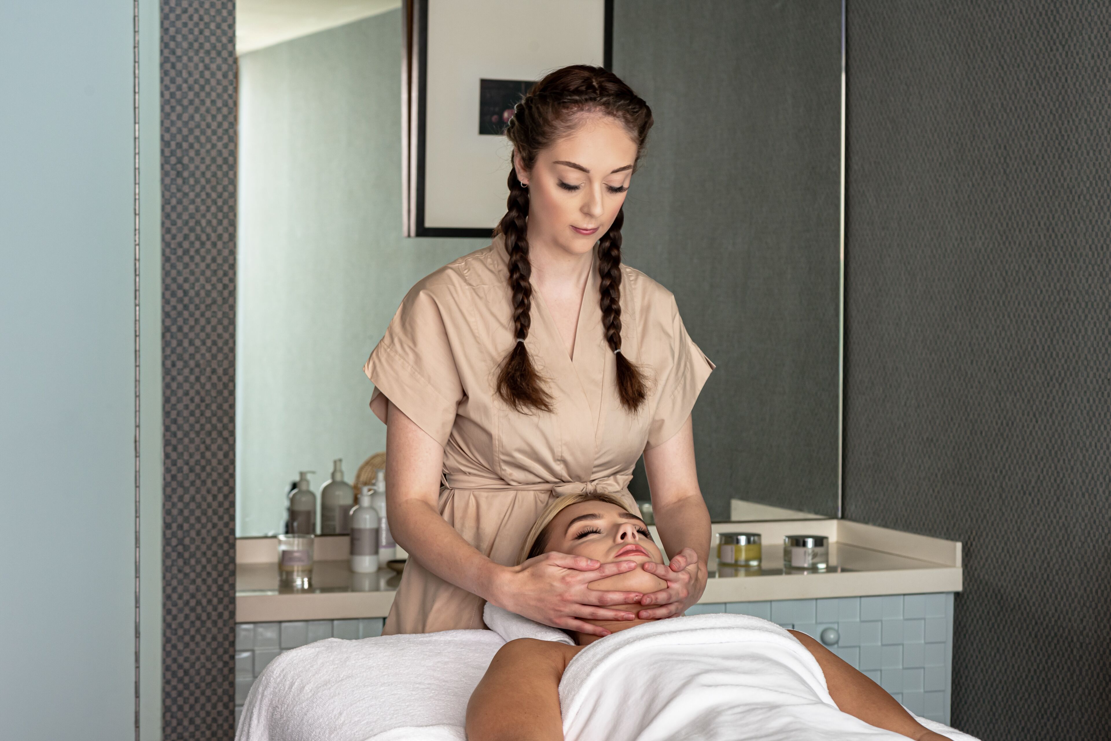 A person receiving a facial treatment in a room with a mirror, a sink, and toiletries on the counter.