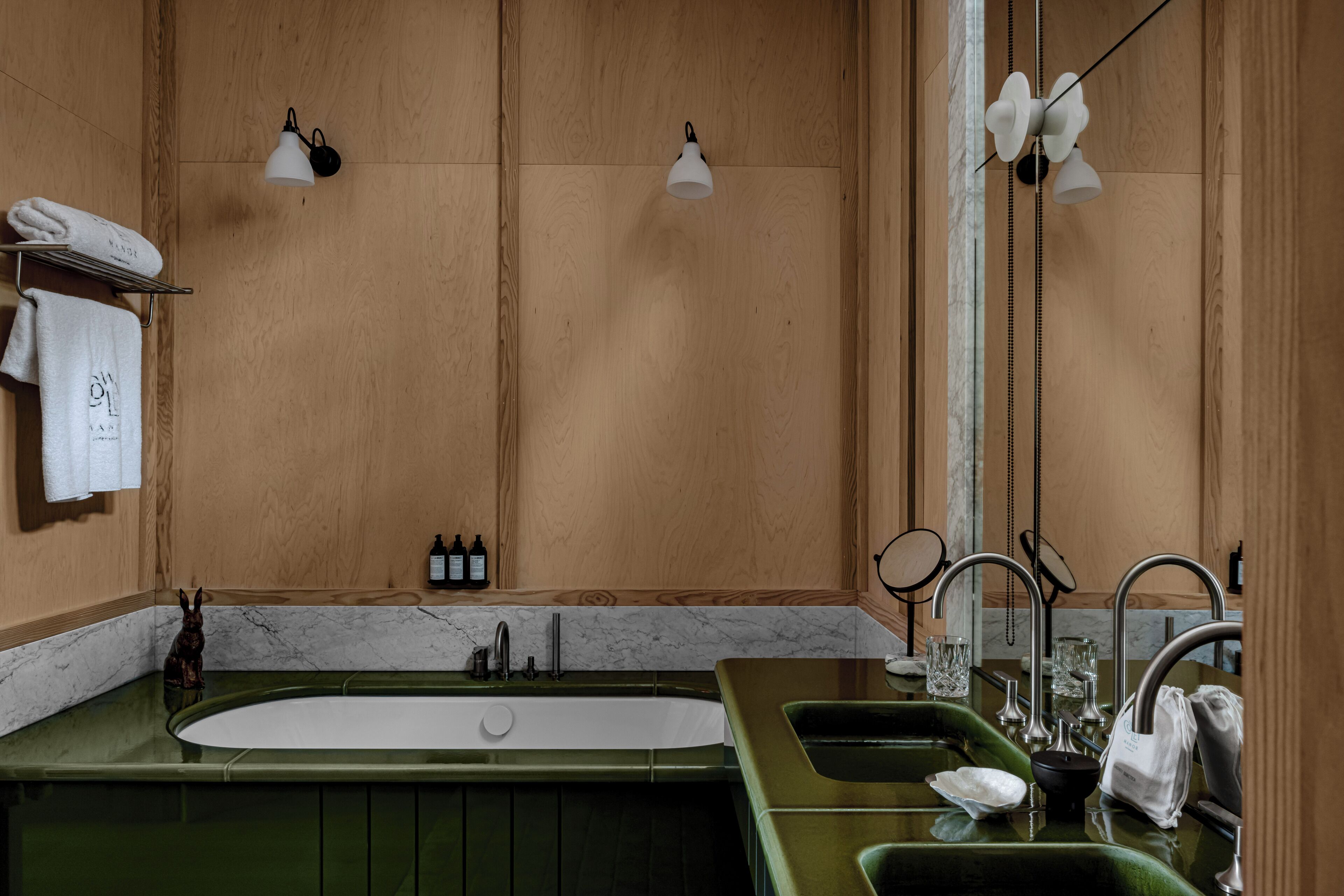 A bathroom with a green vanity, a large bathtub, two sinks, and a mirror. There are towels on a rack and a bottle of shampoo on the counter.