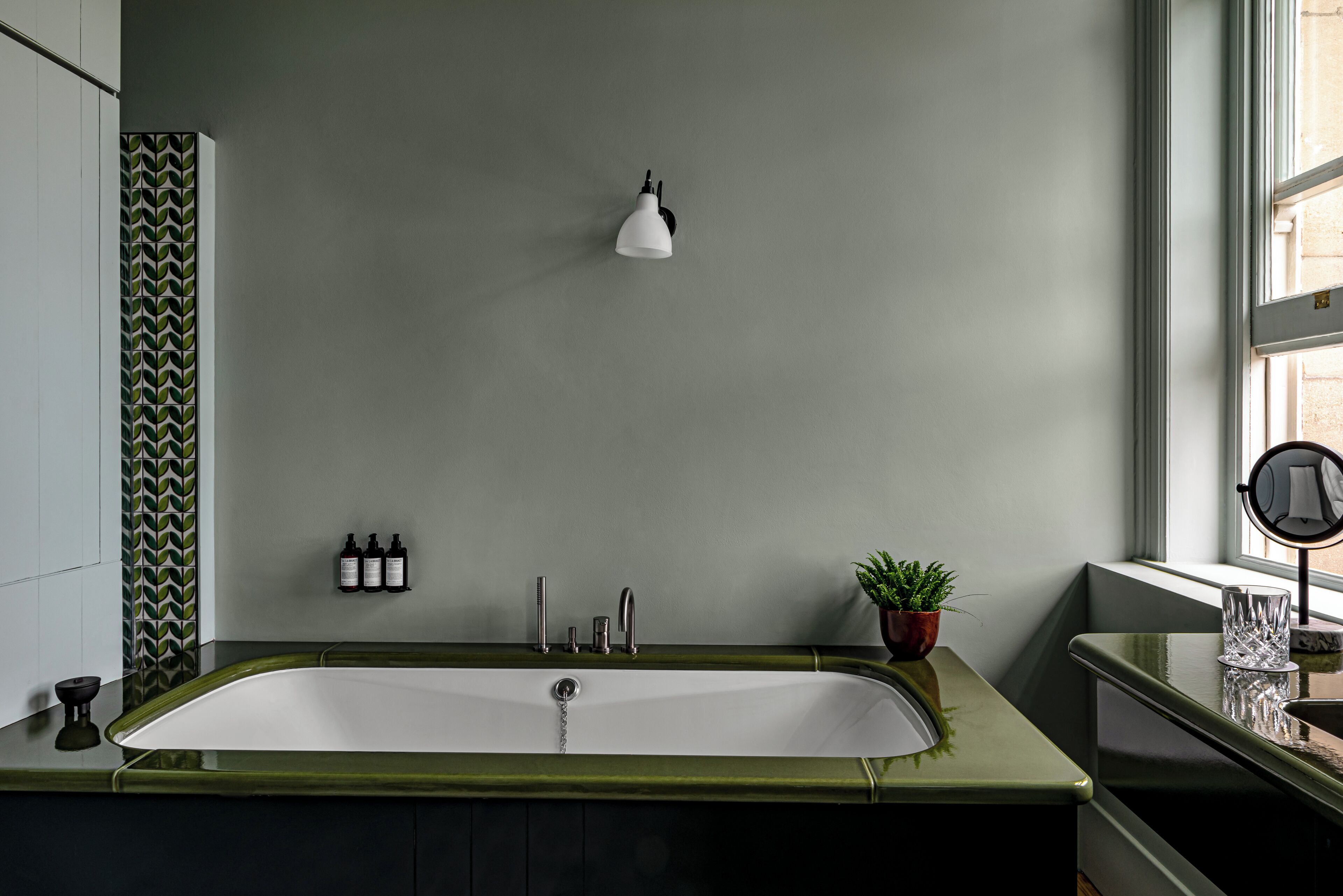 Un baño moderno con un mueble de tocador verde, una bañera blanca, una lámpara de pared, una planta en maceta y un espejo.