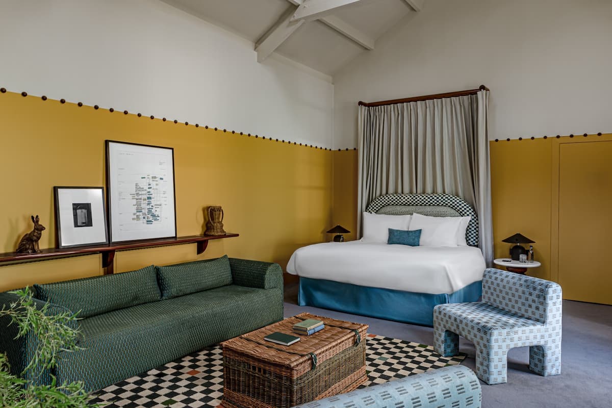 A room with a bed, a green sofa, a small chair, a wicker coffee table, and a checkered floor.