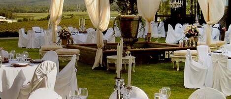 An outdoor wedding reception with round tables set with white tablecloths, wicker chargers, and glassware.
