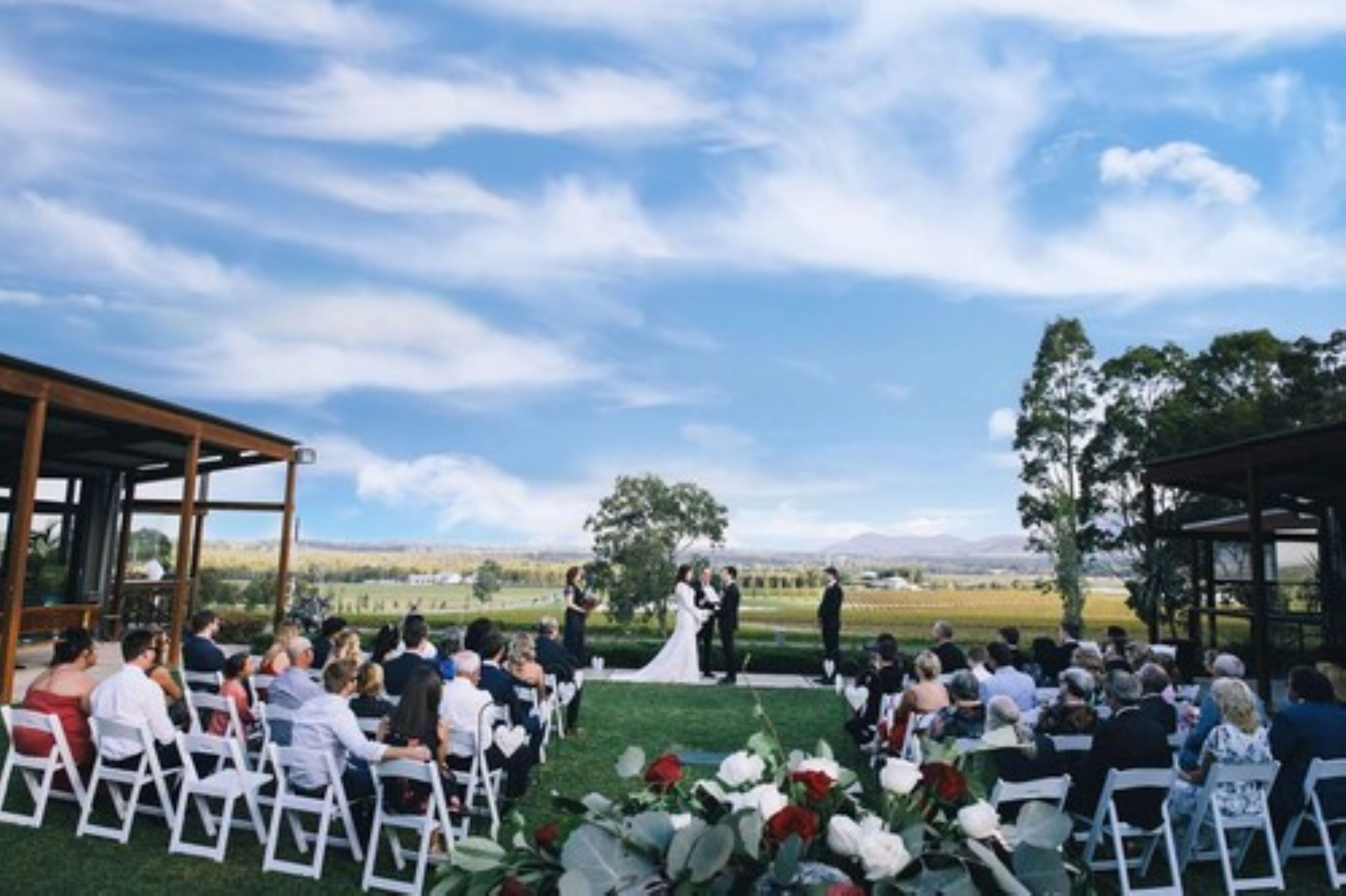 Una ceremonia de boda al aire libre con invitados sentados en sillas blancas, una pareja vestida de boda en el centro, y un cielo despejado.