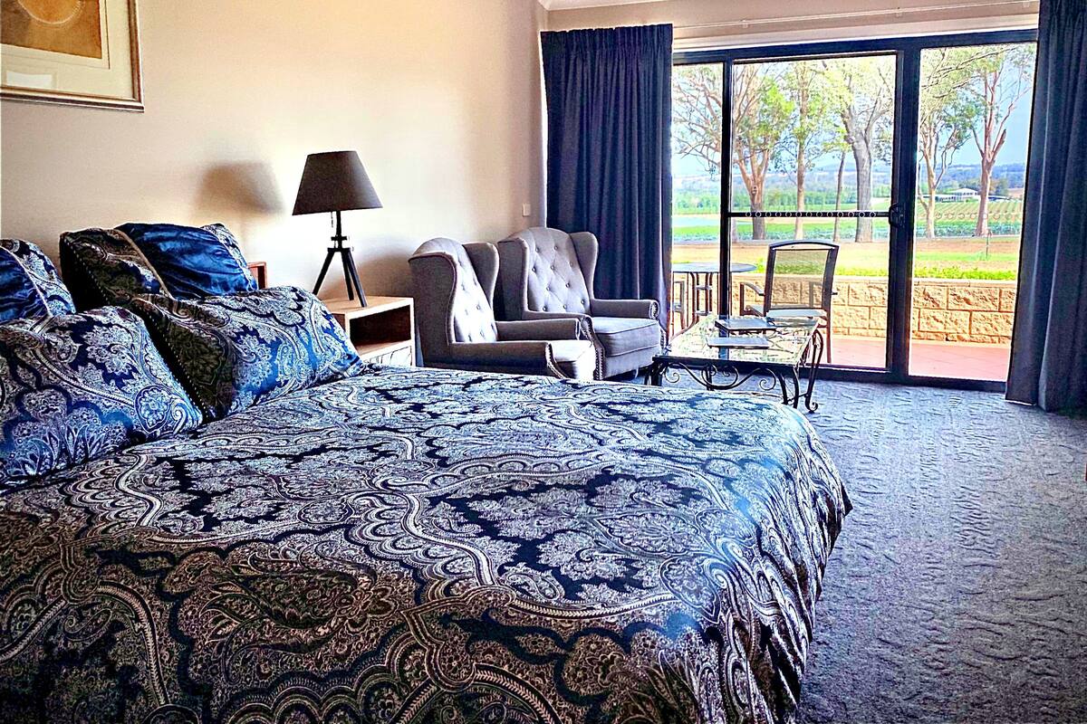 A hotel room with a large bed, a chair, a small table, and a view of the outdoors through a large window.