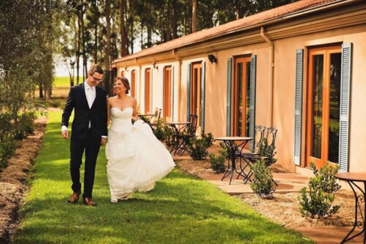 A couple walking on a grassy path beside a building with multiple windows and doors, outdoor seating, and small plants.