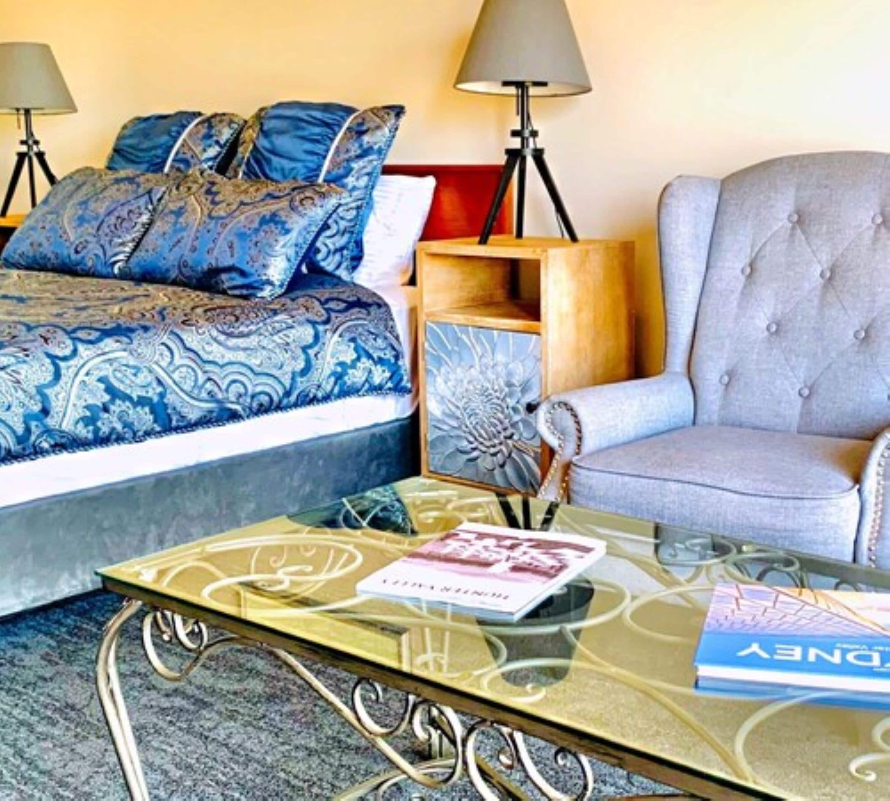 A hotel room with a bed, a grey armchair, a glass coffee table with a book, and two lamps.