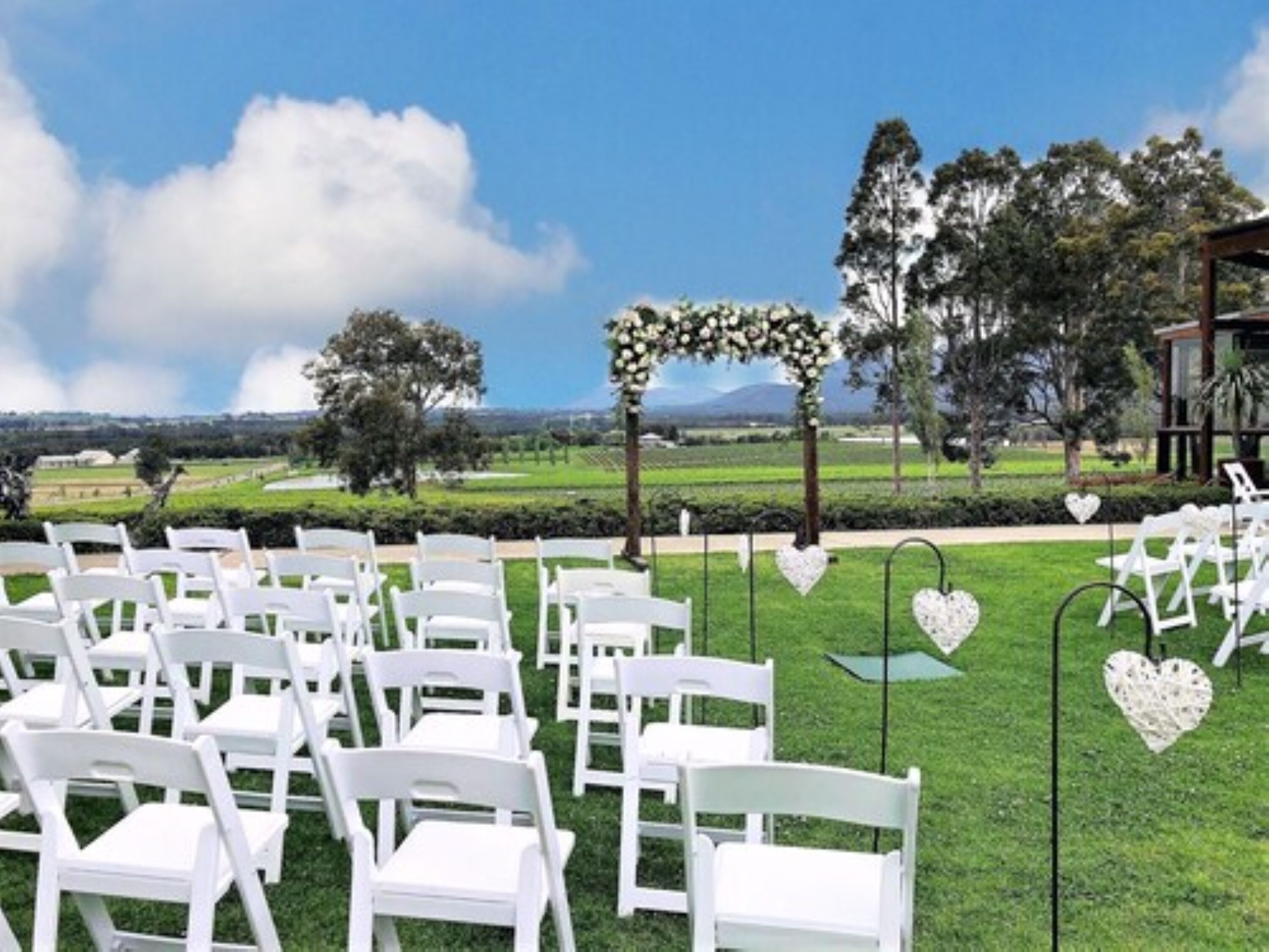 Montaje para una ceremonia de boda al aire libre, con sillas blancas dispuestas, un arco floral y decoraciones en forma de corazón en postes.