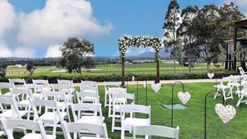 Montaje para una ceremonia de boda al aire libre, con sillas blancas dispuestas, un arco floral y decoraciones en forma de corazón en postes.