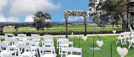 A wedding ceremony setup with white chairs arranged outdoors, a floral arch, and heart-shaped decorations on poles.