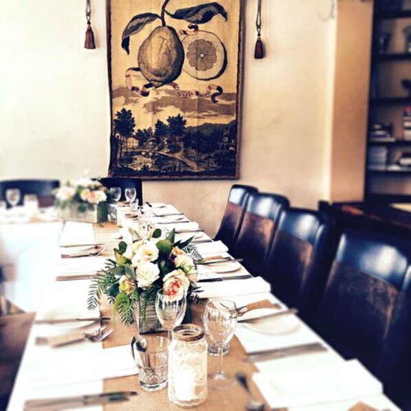 Una larga mesa de comedor vestida con manteles blancos, platos ordenados y copas. Cada mesa tiene centros de mesa con flores, y la habitación tiene un ambiente rústico con sillas de madera y un cuadro grande en la pared.